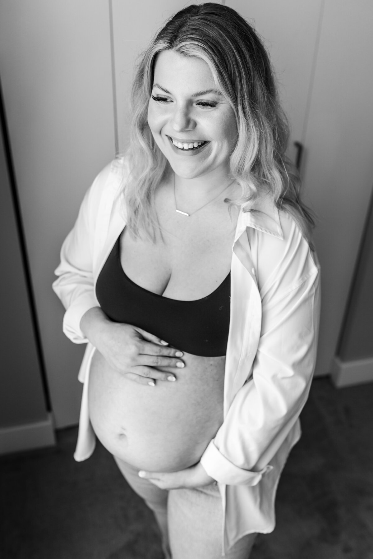 A pregnant woman stands in a bright room, smiling and looking down while holding her baby bump, dressed in a black bra and white shirt.