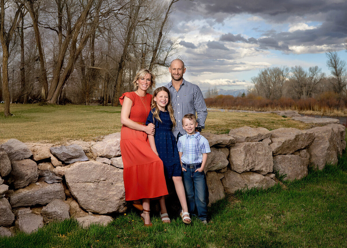 A family of four smiles warmly beside a rustic stone wall, with natural trees and a dramatic sky adding charm to the outdoor setting.