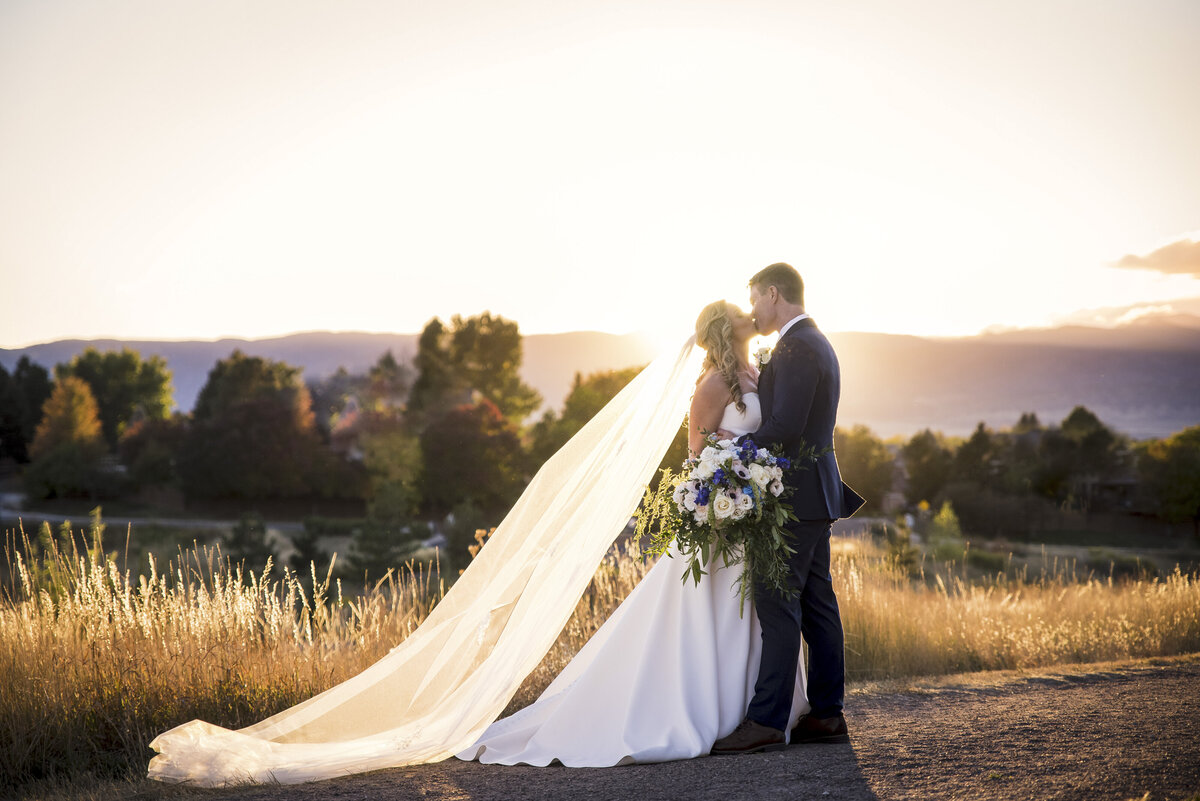 A bride and groom share a kiss at golden hour as the veil floats in the wind.