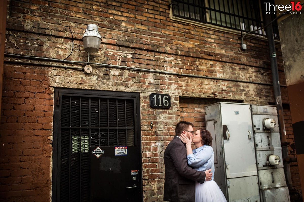 Engaged couple share a smooch in front of an old brick building