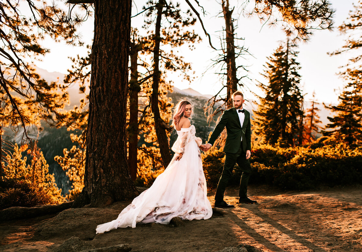 Couples Photography, Man in a blue suit holding hands of a woman in a wedding dress with light pink hair, in the forest