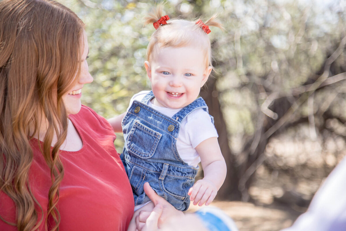 Happy toddler in denim dress being held by mom in a red shirt