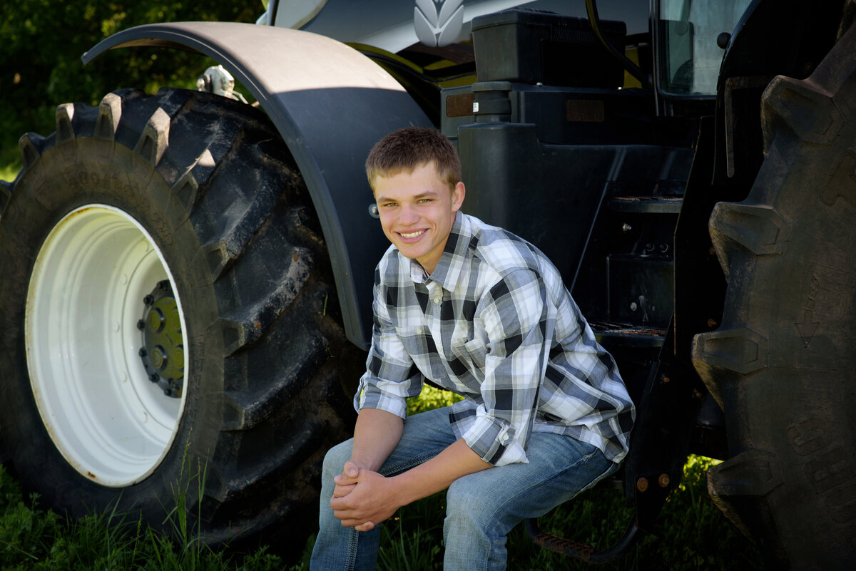 Luxemburg Casco high school senior boy wearing a black and white plaid button down shirt and jeans by a black New Holland tractor in farm field at his home near Green Bay, Wisconsin.