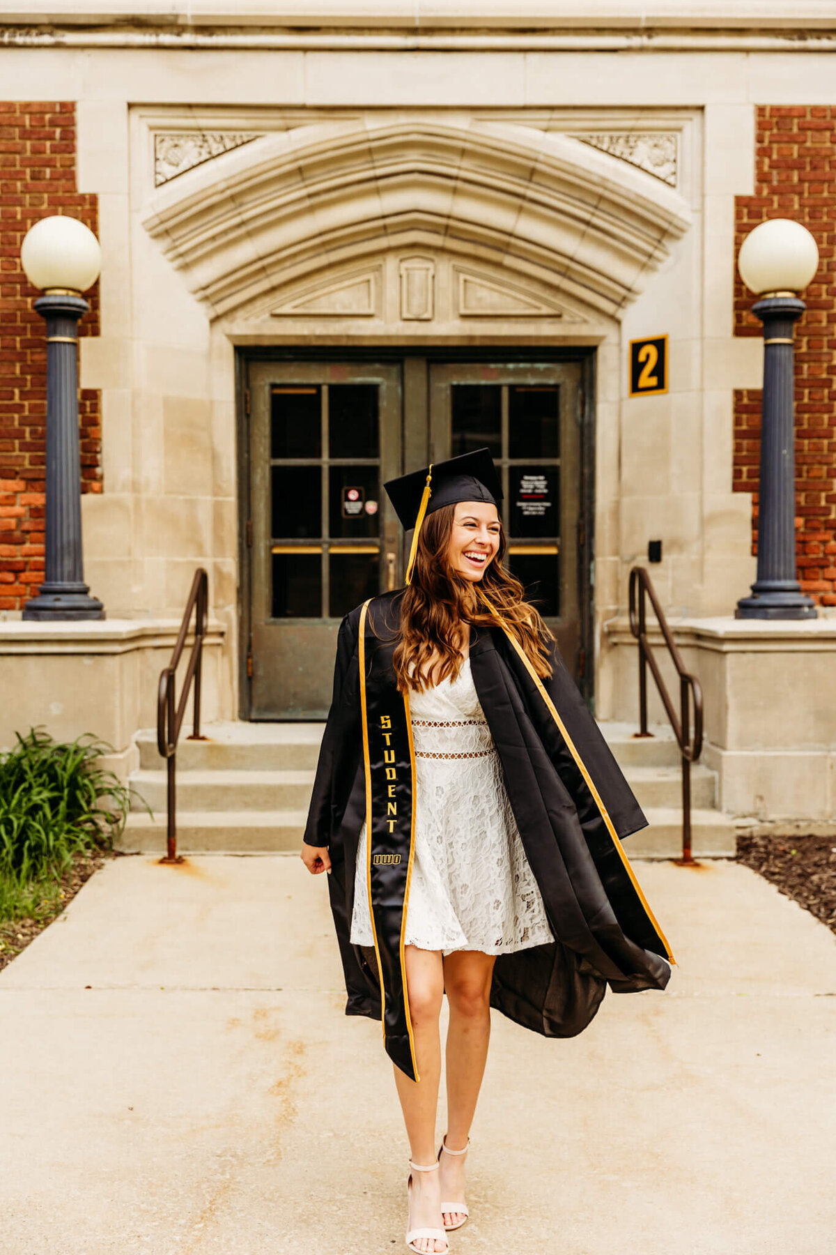 Pretty University of Wisconsin Oshkosh graduate in a white dress wearing her cap and gown laughing as she walks in front of a building during her college graduation photo session