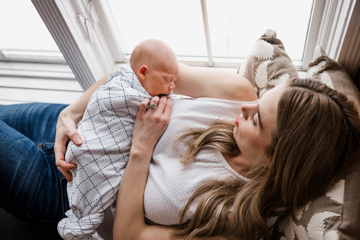 A mother lies back on a window seat, cradling her newborn baby who is swaddled in a patterned blanket, as she gazes down lovingly at the child.