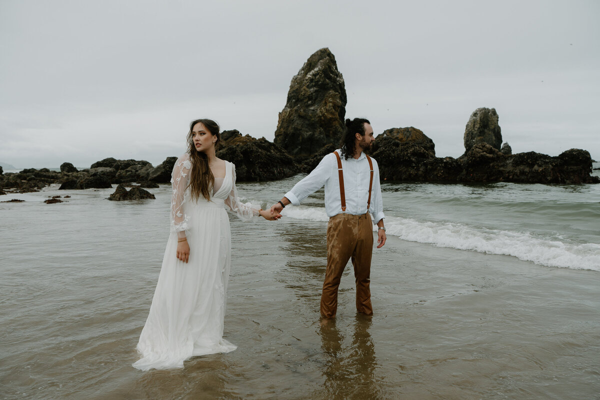 A couple stands in the ocean in front of black rocks at their Cannon Beach elopement. 