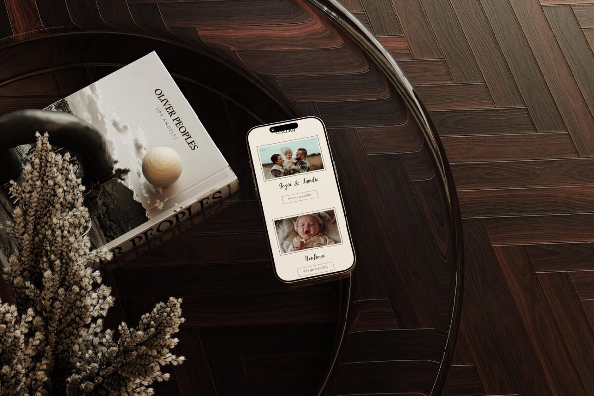 Smartphone placed on a dark wood coffee table next to a photography book and dried floral arrangement, displaying a mobile gallery page with family and newborn portraits.