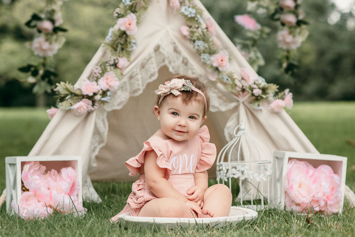 Cute baby girl smiling at camera wearing pink ruffle  romper sitting outside in grassy area with teepee decorated in florals
