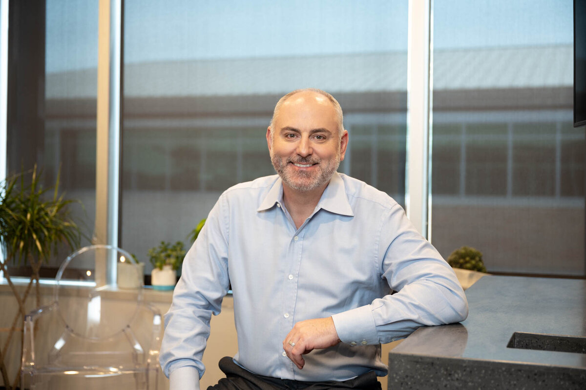 a headshot of a man  sitting in front of a desk with his arm on it and the background is a view of a building