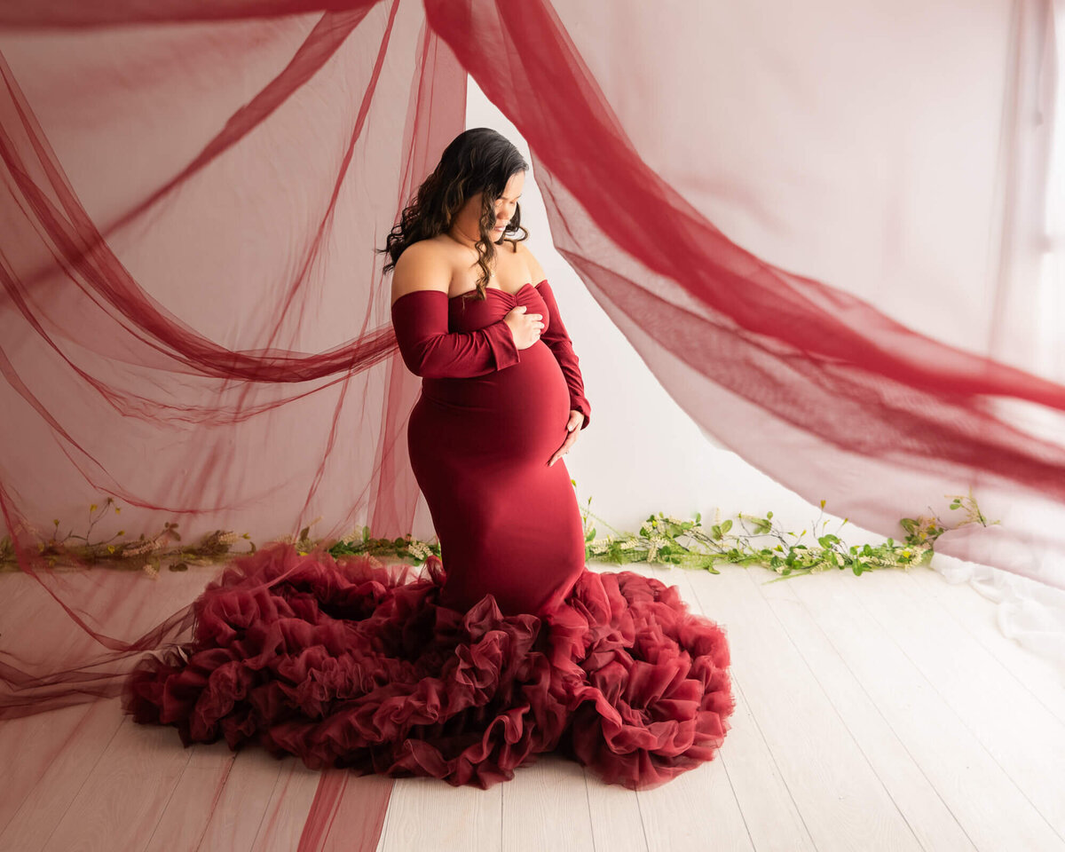 A pregnant woman in a stunning red gown poses gracefully for a photo, showcasing her beautiful baby bump.