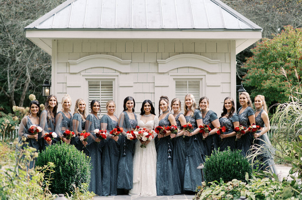 Bride and bridesmaids standing in front of cottage holding red bouquets. 