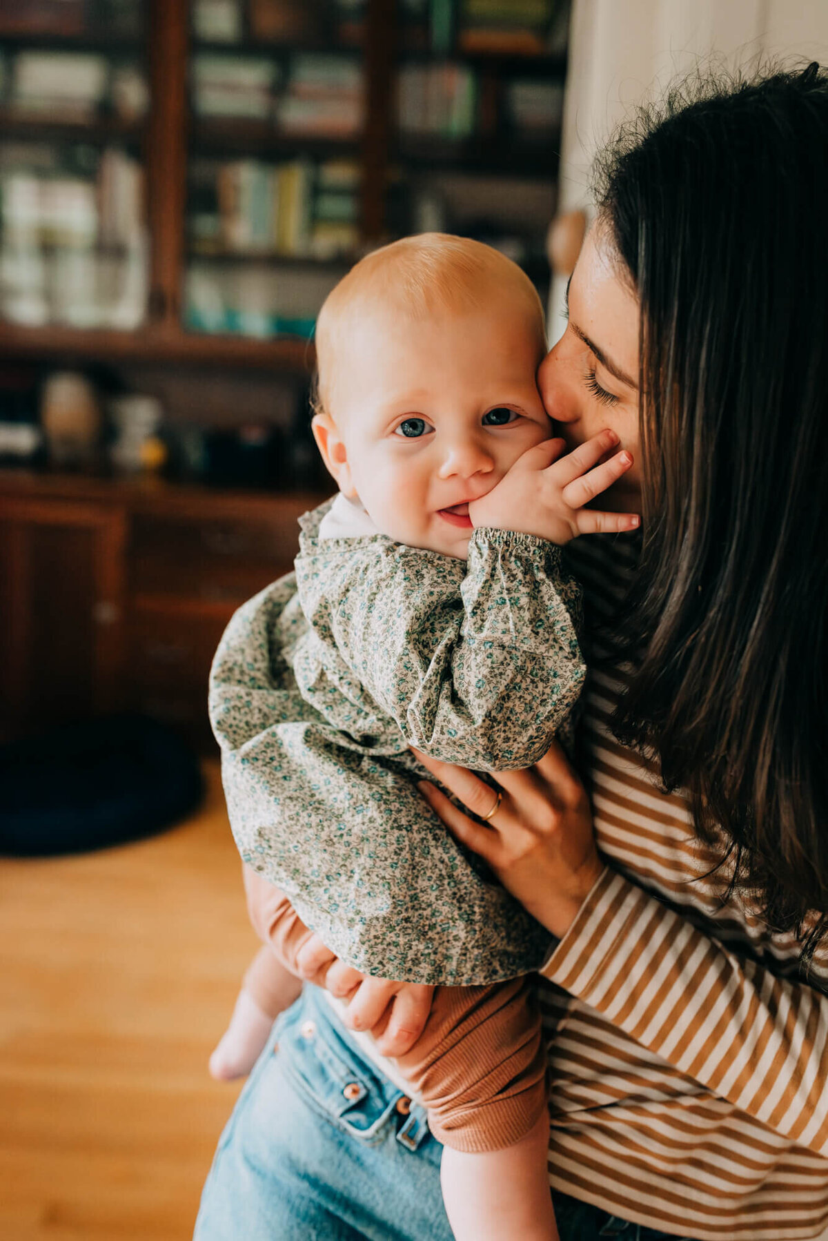 A mother holds her daughter in their kitchen while her daughter sucks her thumb and the mother gives the daughter a kiss during their photoshoot with Allison Wolf, Botson Family Photographer
