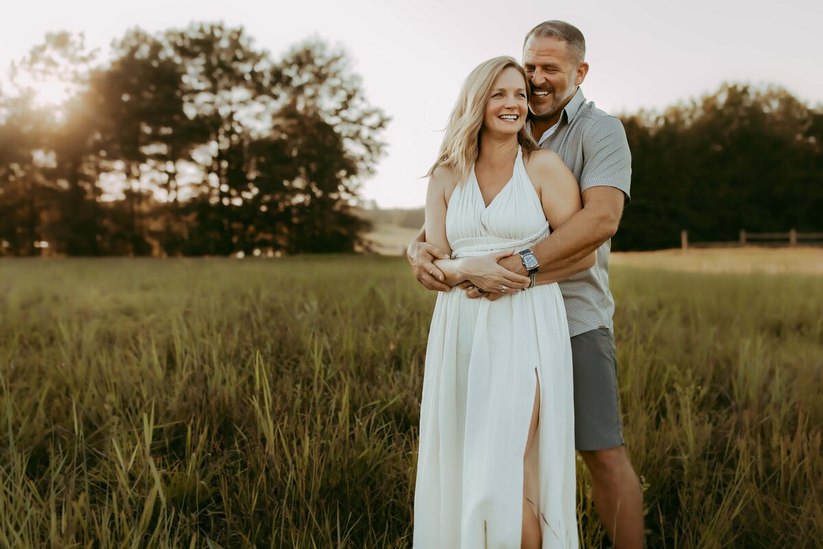 A couple stands in a grassy field at sunset, smiling and embracing. The woman wears a white dress and the man is in a gray shirt and shorts. Trees are visible in the background, capturing a perfect moment of fire family photography by a talented Warner Robins family photographer.