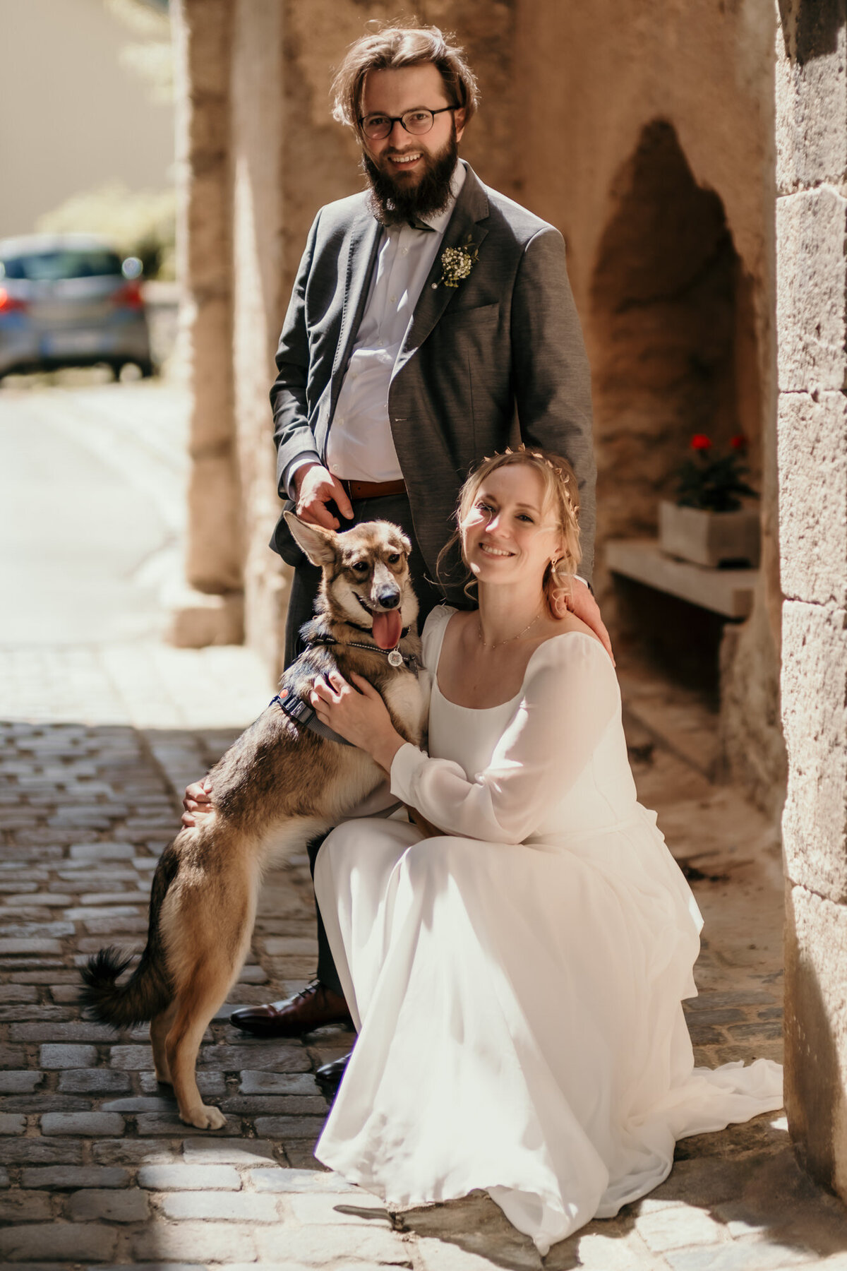 Der Bräutigam steht hinter der knieenden Braut, ihr Hund steht mit den Vorderbeinen auf ihr.