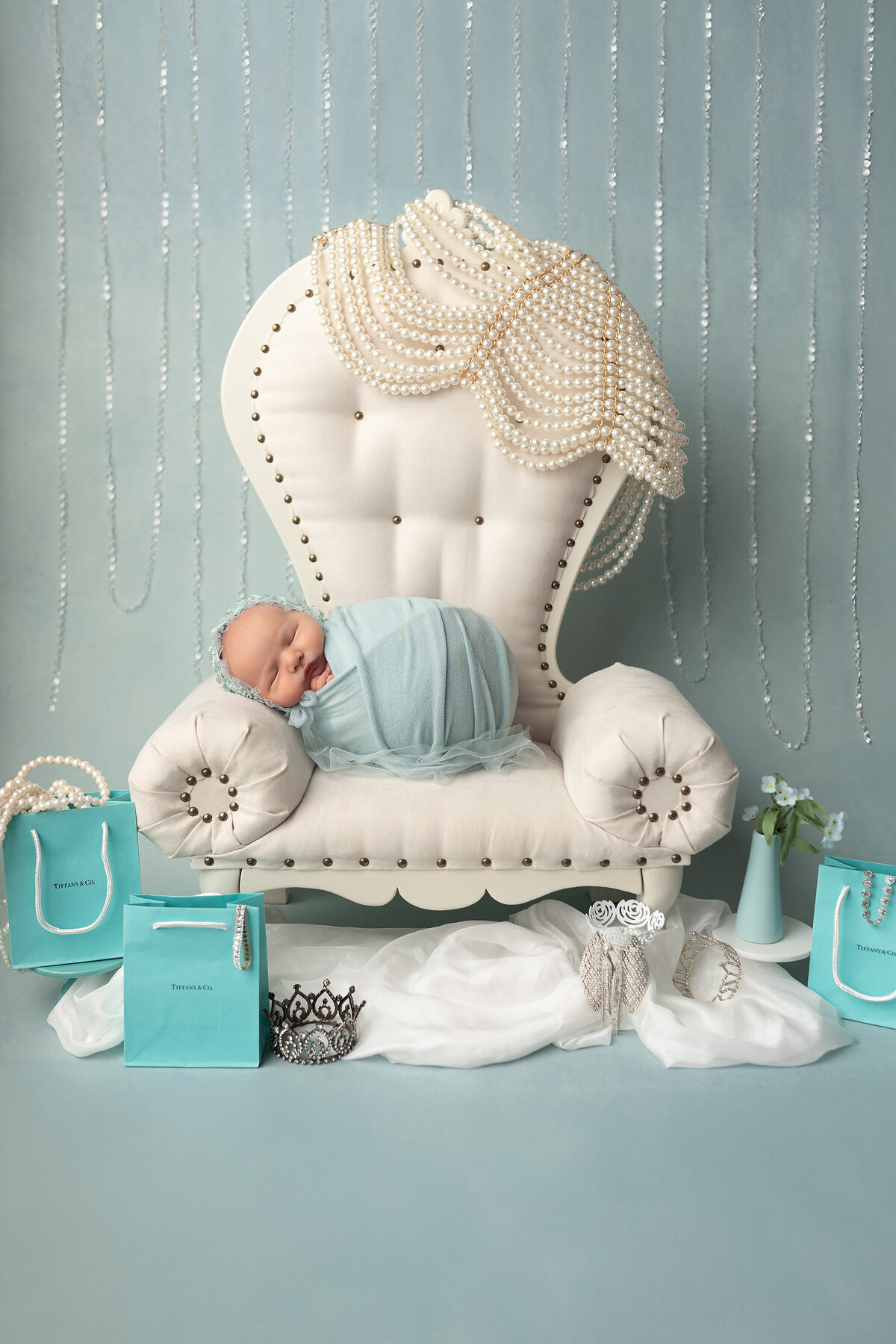 Elegant newborn wrapped in Tiffany blue, posed on a cream throne with pearls, tiara, and Tiffany bags during luxury newborn pictures session in Dallas.