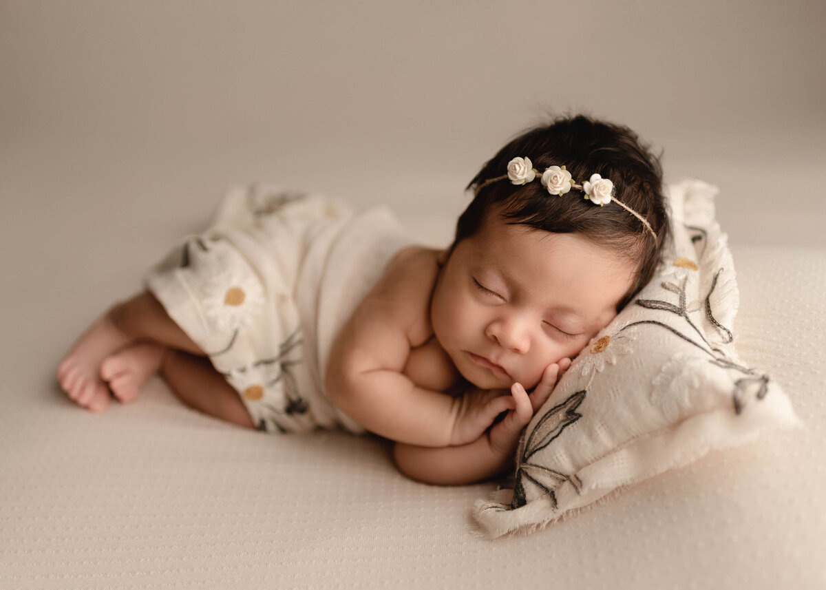 Newborn baby girl laying on side with delicate rose petal headband on cream with sunflower embroidered pillow and blanket