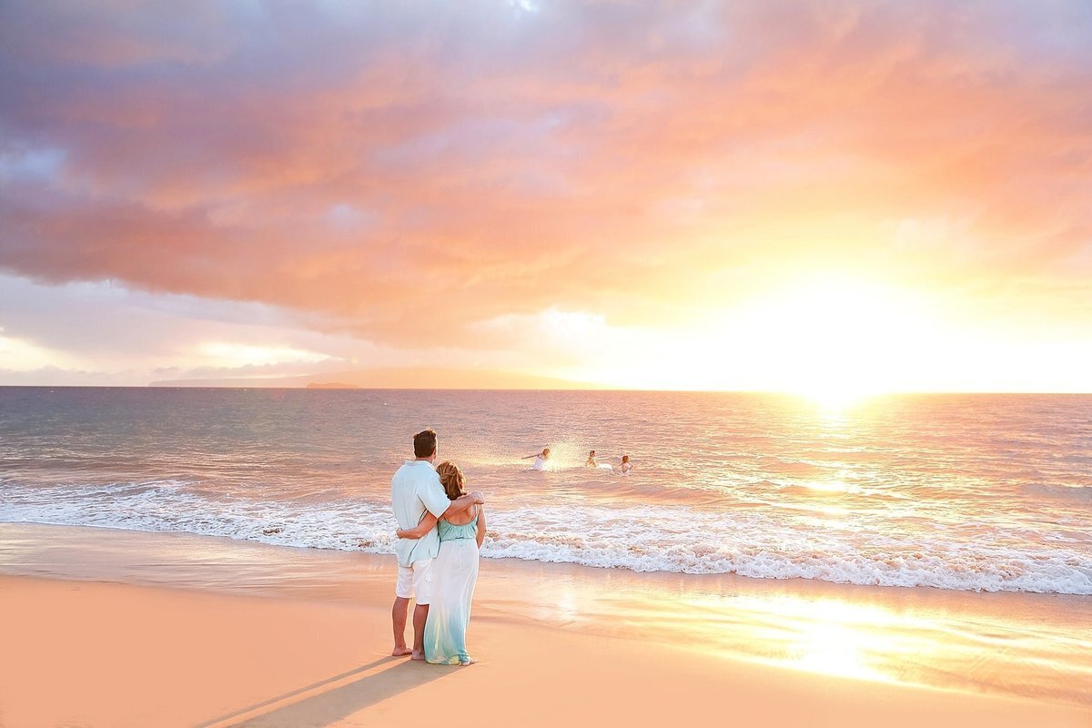 Teen girls splash in the ocean in Maui as their parents watch from the beach with arms wrapped around one another at sunset with Love + Water