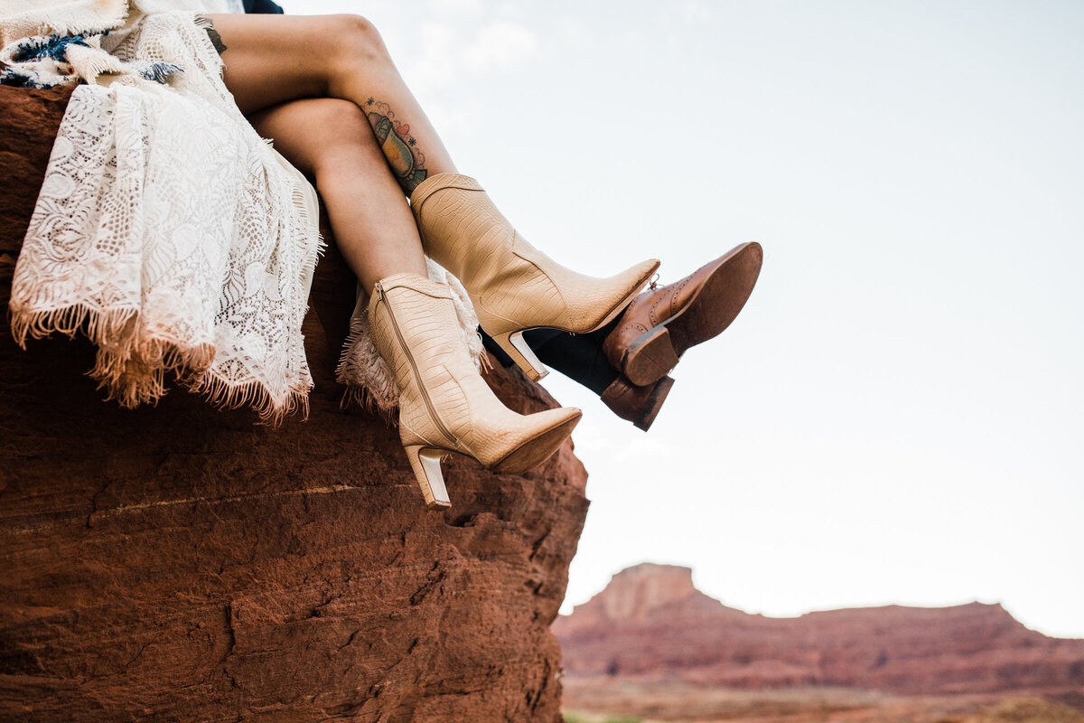 the boho bride sits in a slot canyon and laces her boots. her lace dress fans out beside her.