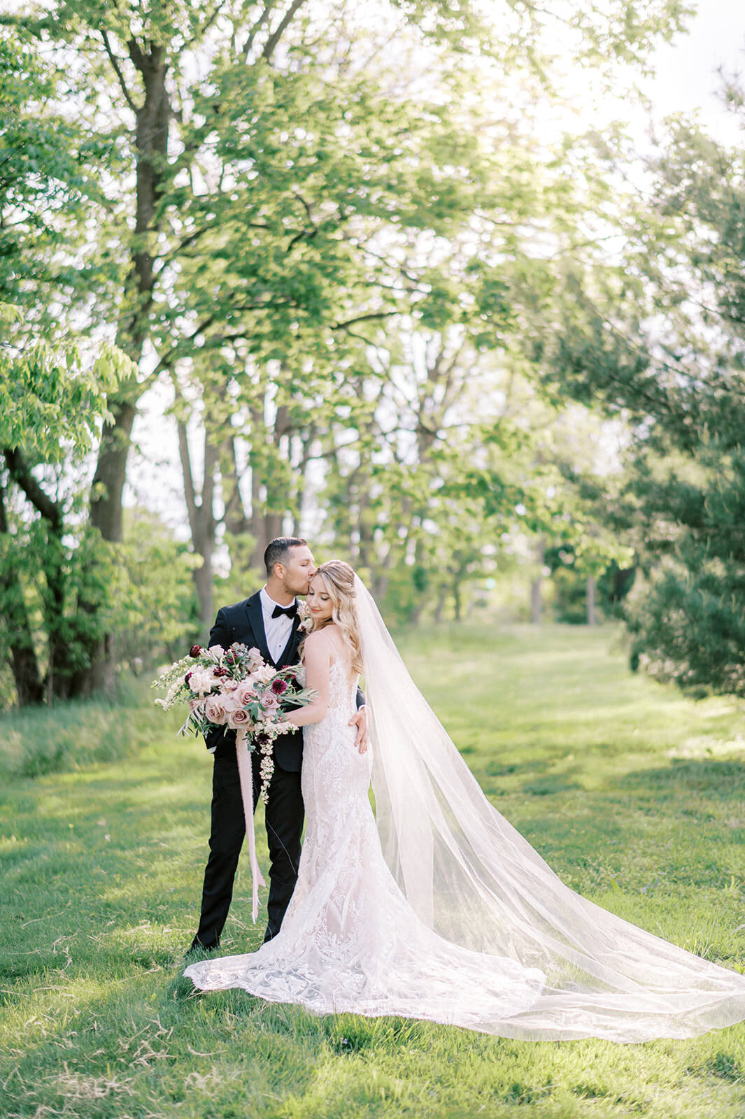 A groom in a  classic black tux kissing his bride with mauve florals