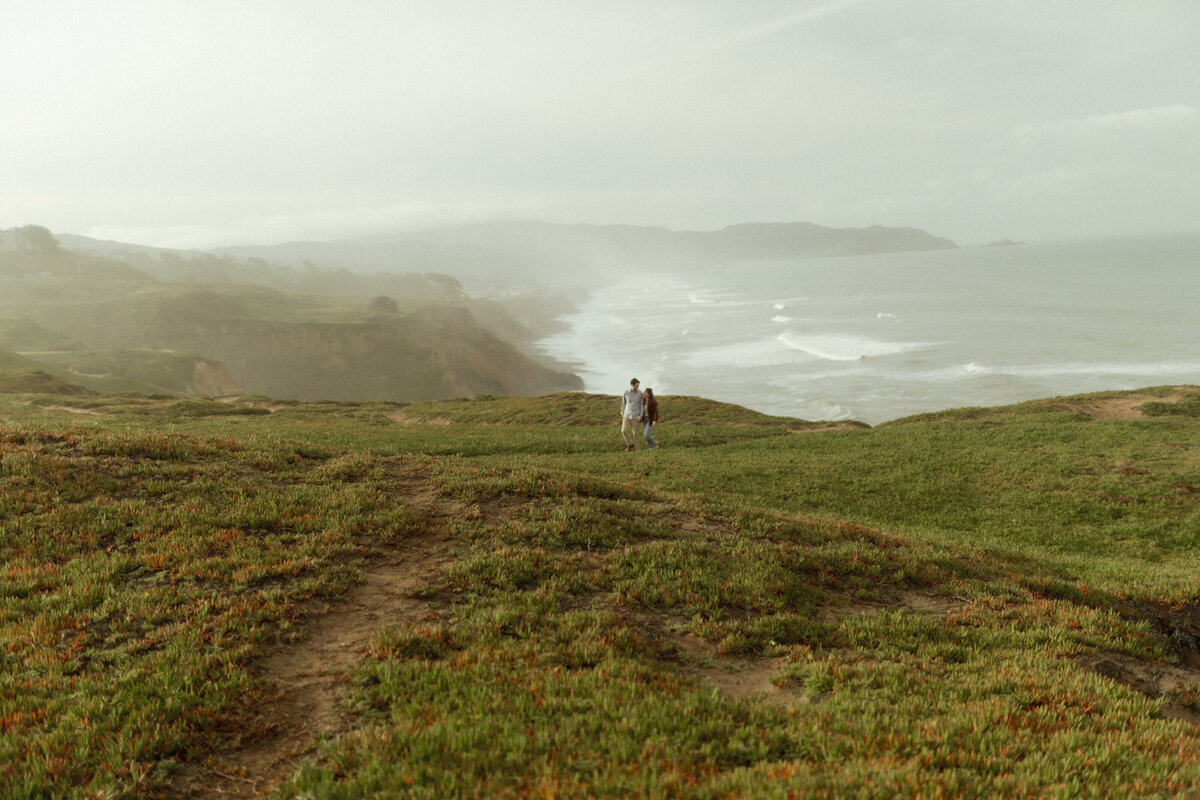 Guy and girl walking during engagement session on California coast cliffs with greenery and fog coming in