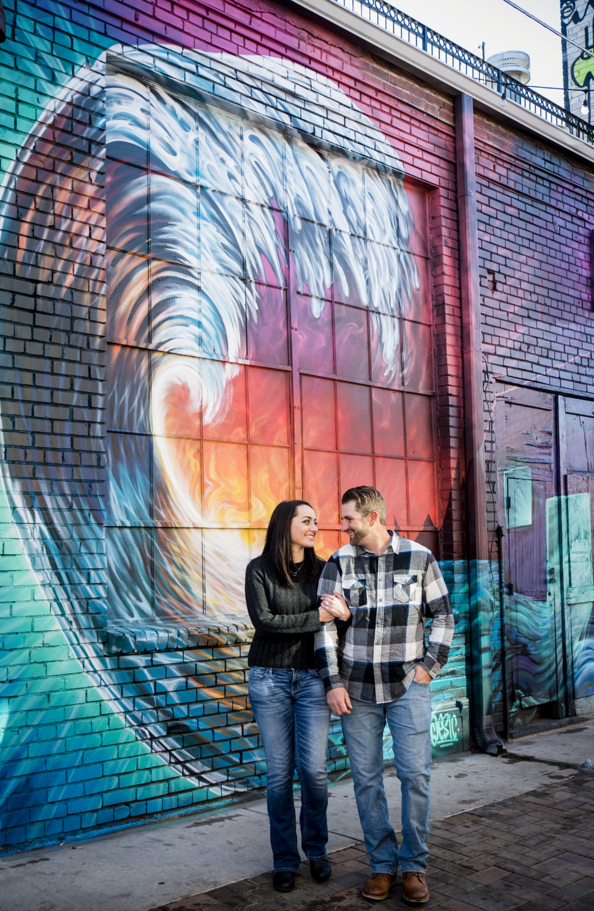 A woman holds her fiancé's arm as they stand in front of a colorful mural for their downtown Denver engagement session.