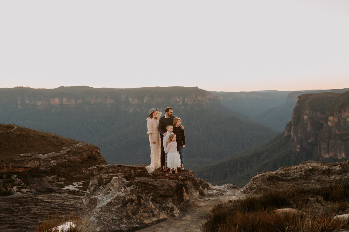 Family of five standing together on a mountain cliff in the Blue Mountains during golden hour, looking out over the scenic valley for a timeless family portrait.
