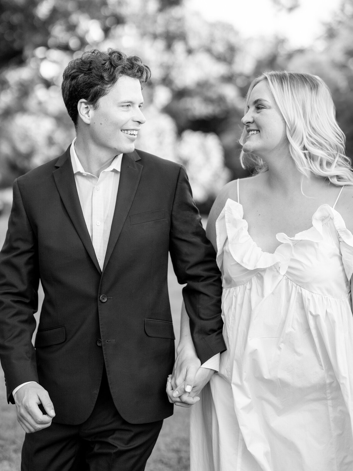 A couple holds hands and walks outdoors, smiling at each other. The man wears a dark suit, and the woman is in a white dress. The background is a blurred natural setting, suggesting a park or garden. The photo is in black and white.