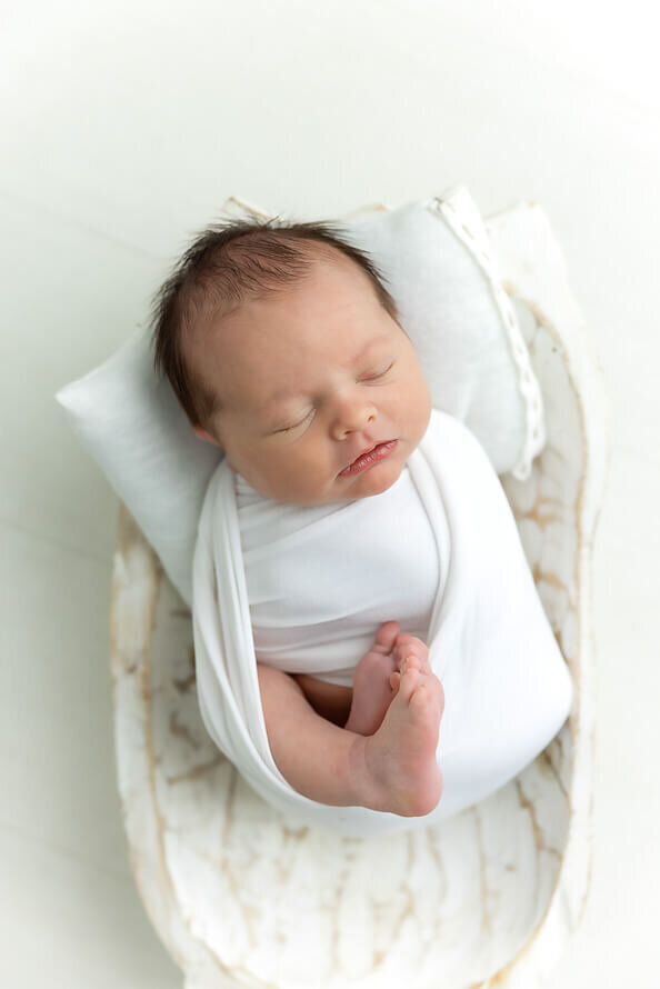 Baby wrapped in white sleeps in a bowl with a pillow.