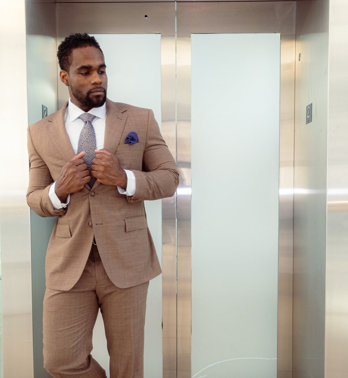 A man posing in a tan suit against an elevator
