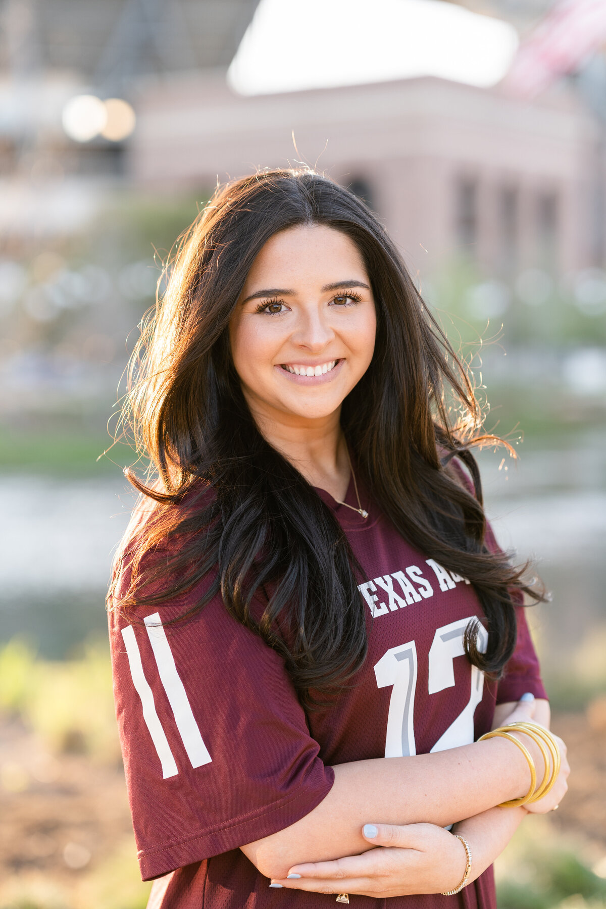 Texas A&M senior girl smiling with arms crossed wearing maroon Aggie jersey in Aggie Park at sunset