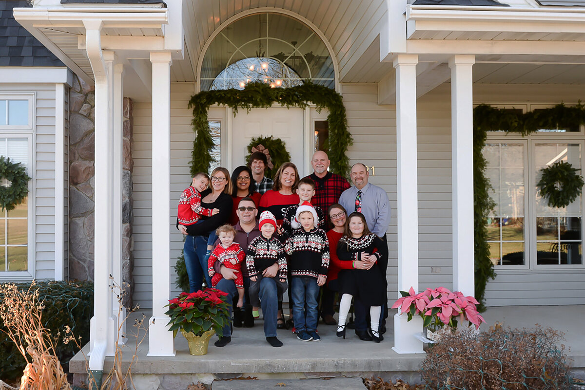 Wearing christmas sweaters, a family sits on their front porch that’s decorated for winter