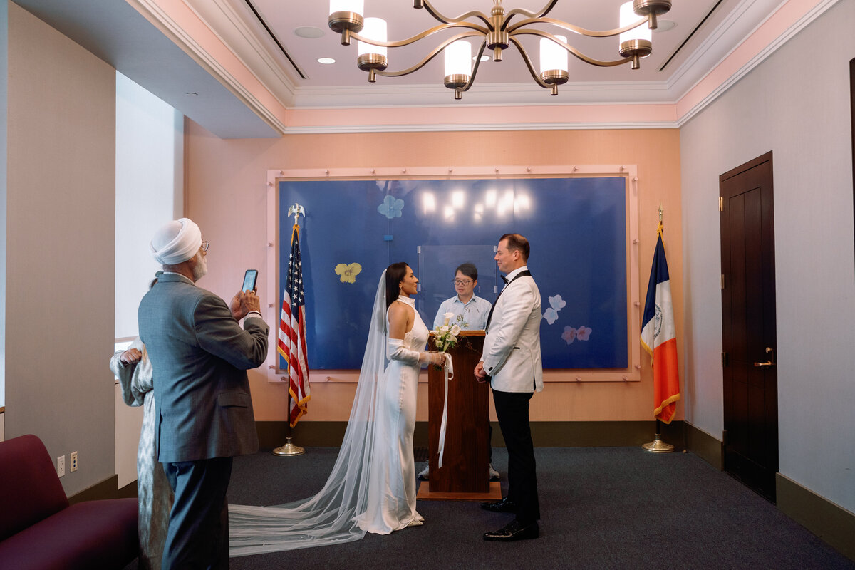 Bride and groom exchange vows during their New York City Hall wedding ceremony, standing before the officiant with family looking on and capturing the moment, photographed by Perry Hancock NYC elopement photographer.