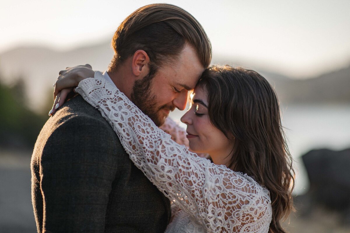 A woman in a lace wedding dress and a man in a tan suit stand at a viewpoint with the Columbia River Gorge as their backdrop