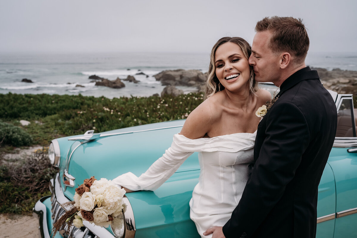 candid feeling moment of couple eloping in monterey coast, blonde latina bride leans on  vintage teal chevy bel air convertable, smiling latina bride in white off shoulder gown holds bouquet, white groom in black suit kisses her ear