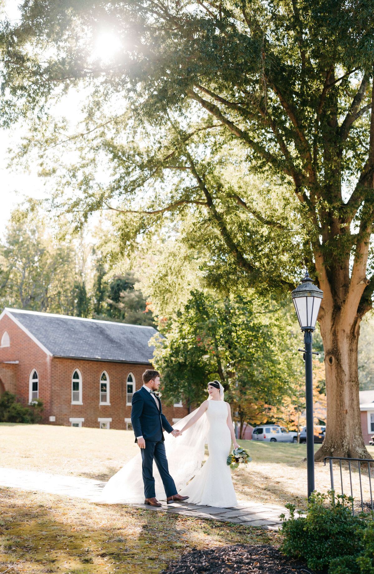 wedding in Richmond va with bride and groom walking down a hill from a wedding chapel while holding hands and being illuminated by the setting sun as they smile at one another as they aproach a old school street lamp leading to a stone path