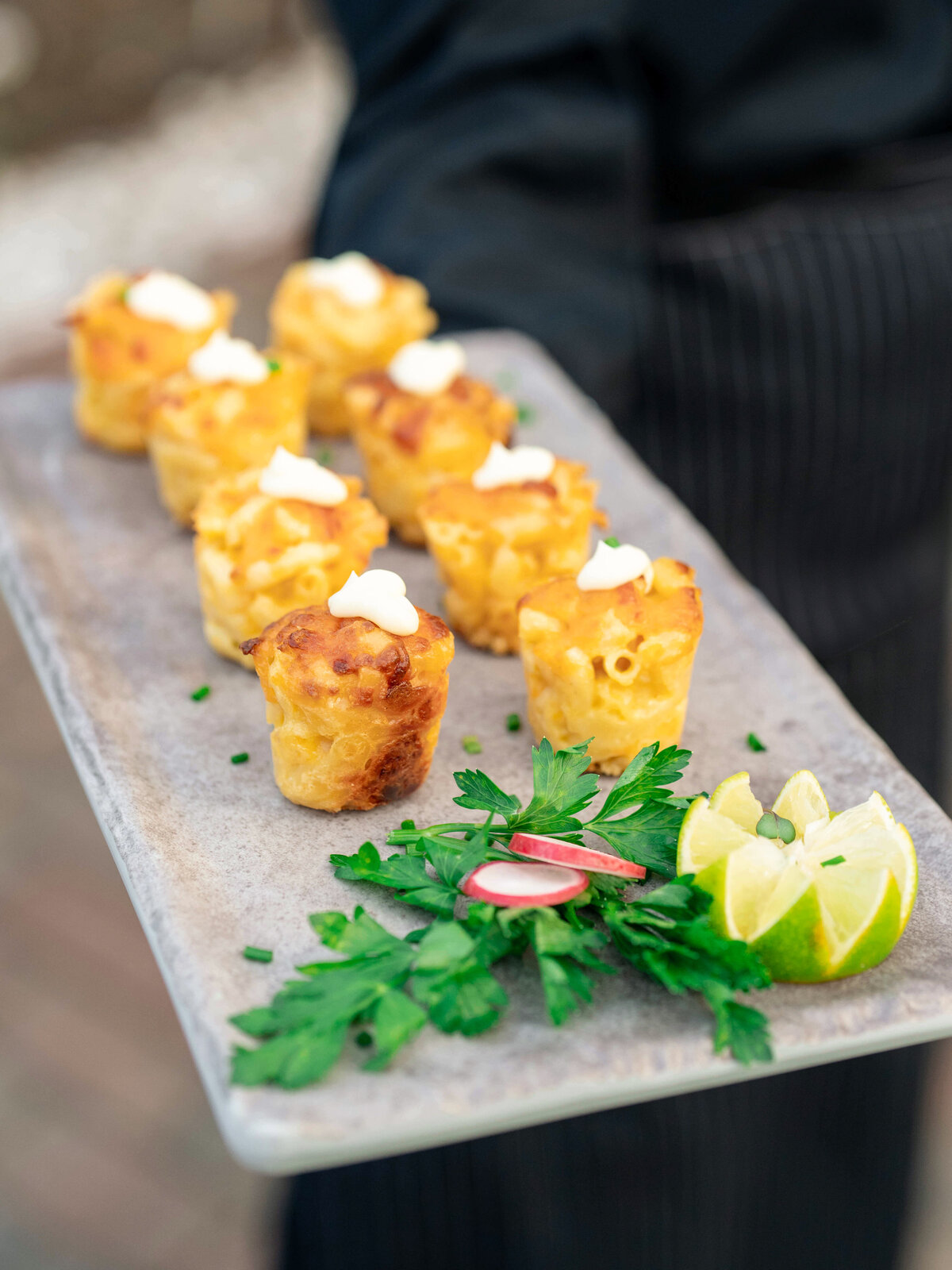 A server holds a tray with several baked macaroni and cheese bites, each topped with a dollop of white sauce. Garnished with parsley, lime wedges, and radish slices on the side. The server wears a black outfit with pinstripes.