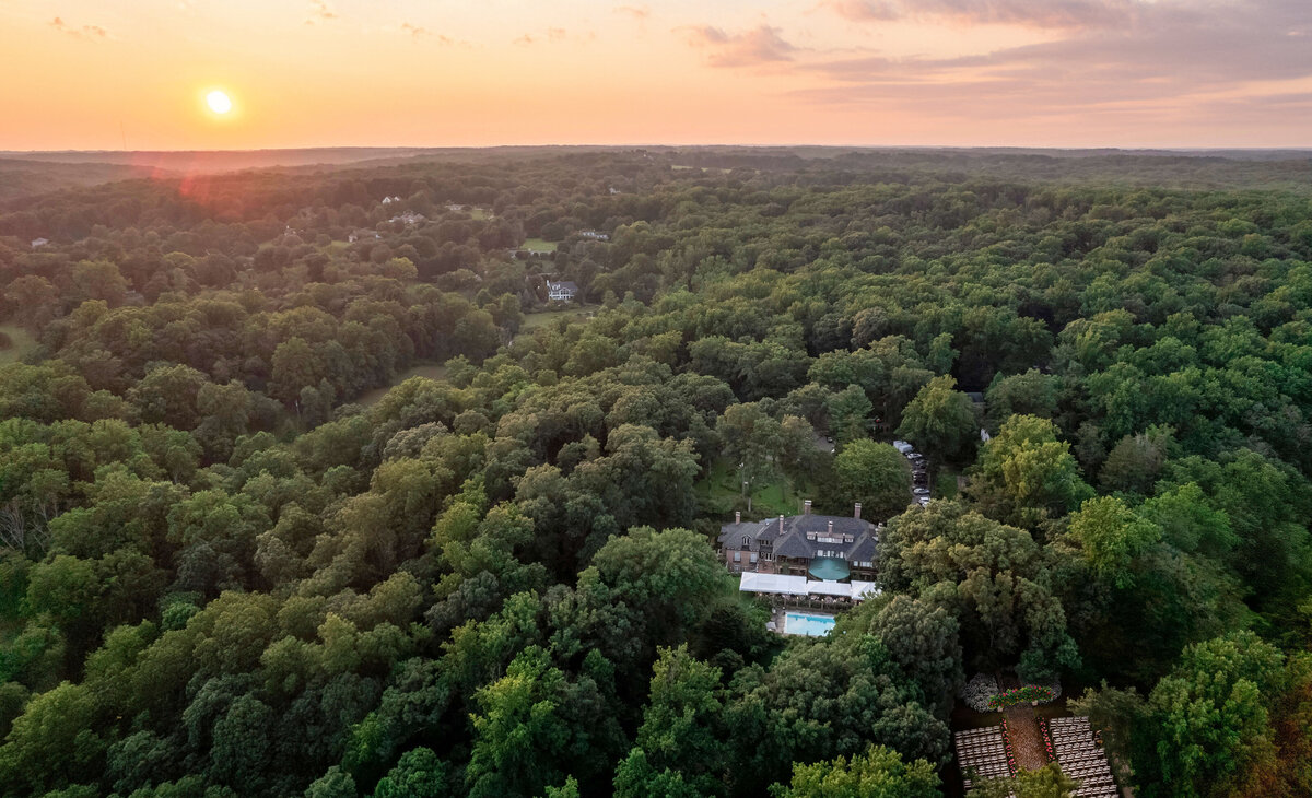 Aerial view of a lush, green forest at sunset, with a sprawling estate featuring multiple buildings nestled among the trees. The sun sets on the horizon, casting a warm glow over the landscape.