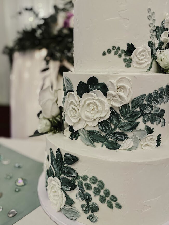 A close up shot of a white cake with concrete textured frosting. Each tier has pallet painted white flowers and greenery.