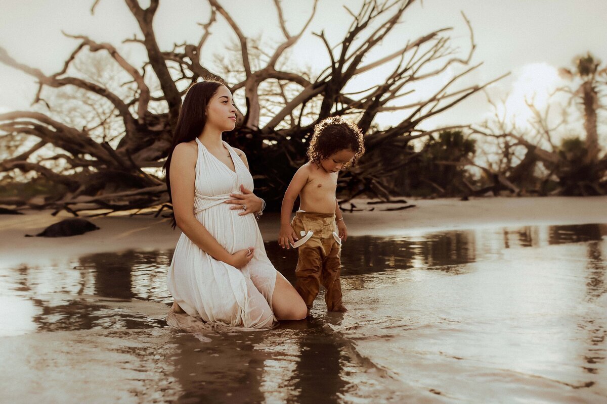 A woman in a white dress kneels by a sandy water's edge, holding her pregnant belly. A child stands beside her, looking down, with driftwood trees in the background.