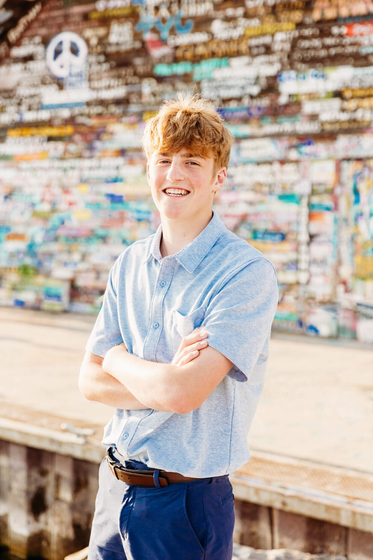 young man in a blue polo crossing his arms and laughing as he stands in front of a multi colored building.