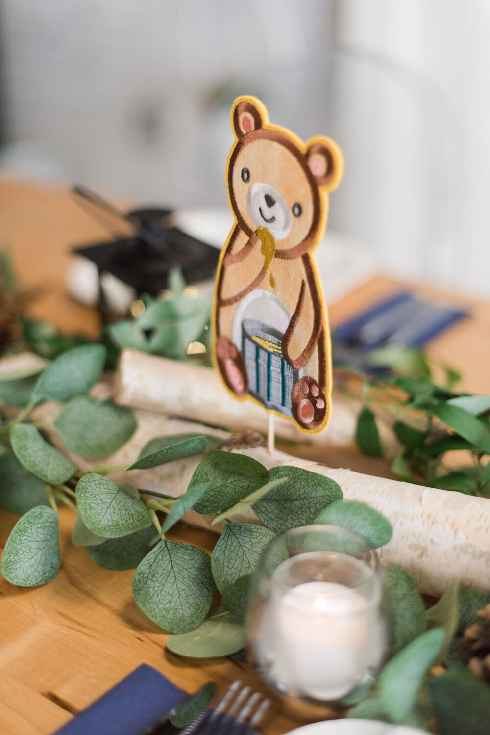 Baby shower centerpiece featuring a bear figurine eating honey, perched on wood logs and surrounded by lush greenery and a vintage lantern.