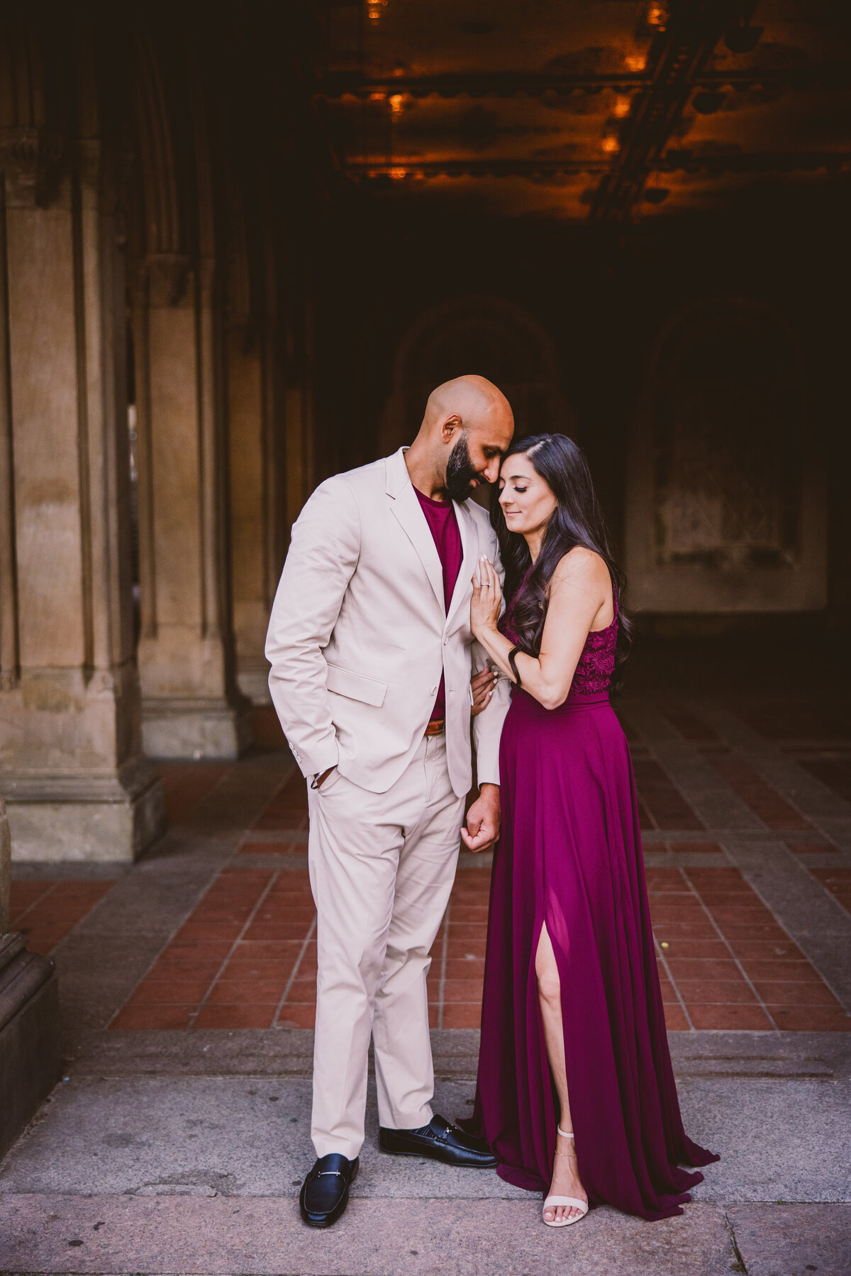 An engaged couple share a moment under the arches at central park bethesda terrace wearing formal red clothing