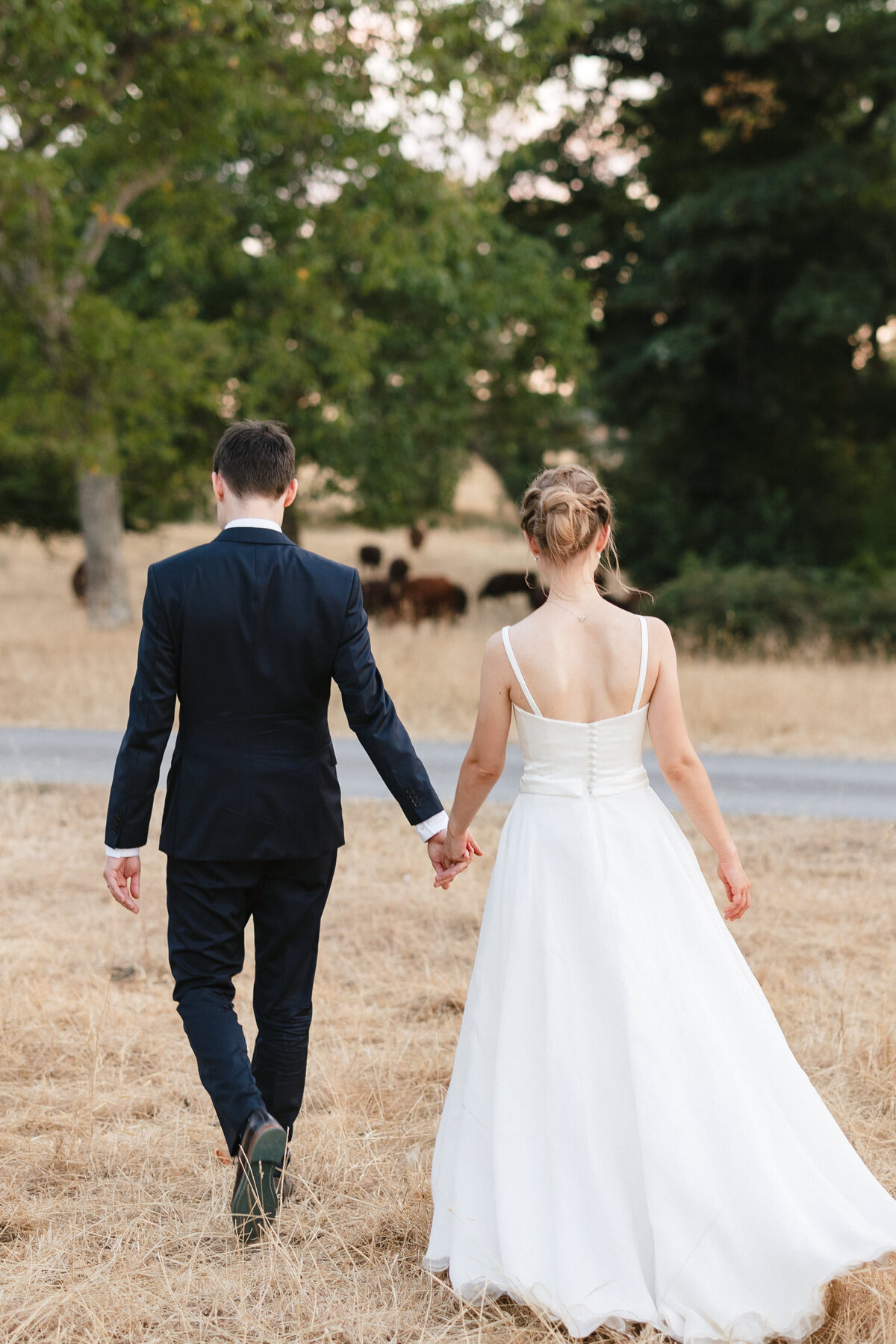 A couple, dressed in a wedding gown and suit, walk hand in hand on a grassy path at Wasing Park. They are facing away from the camera, heading toward trees and a group of grazing animals in the background, embodying the serene beauty of their special day.