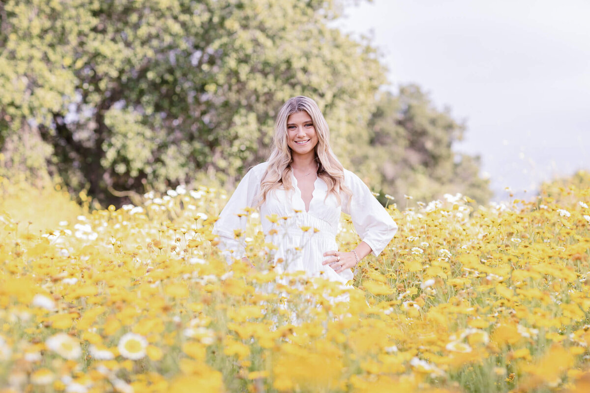 A woman in a white dress stands in a vibrant field of yellow and white flowers, surrounded by lush green trees under a clear sky. She smiles with her hand on her hip.