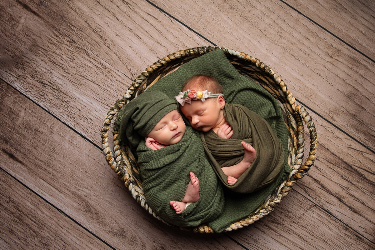 boy girl twins wrapped in green swaddles laying in a basket on a wood floor captured byFort Mill Portrait Photographer