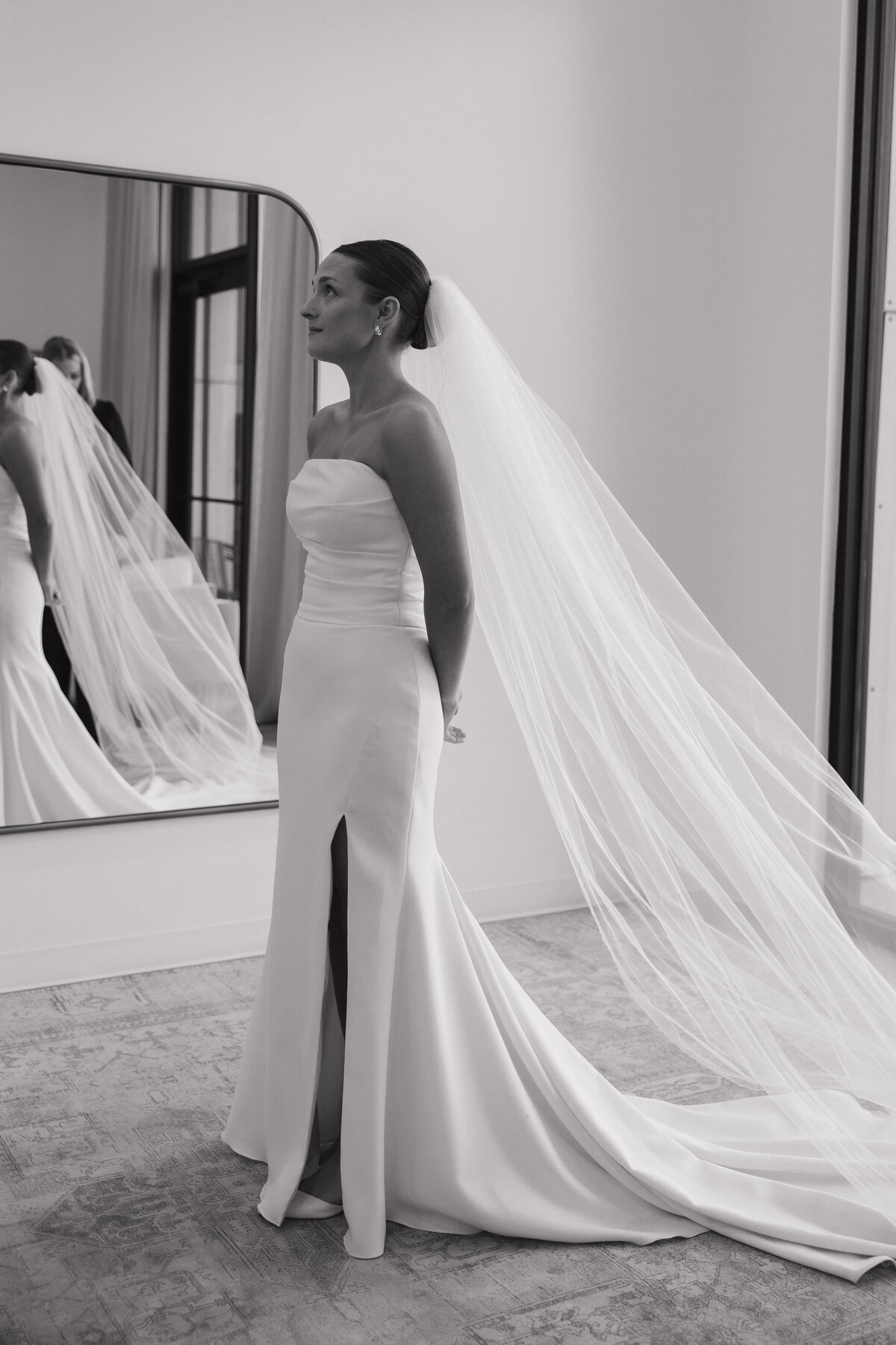 A bride stands looking up toward the ceiling tearfully with her hands laced behind her back with a long cathedral length veil and a fitted white satin dress that is strapless with a thigh high leg slit. There is a half-oval shaped mirror on the wall behind her reflecting her back and veil. She has a slicked back bun underneath her veil and gold hoop earrings