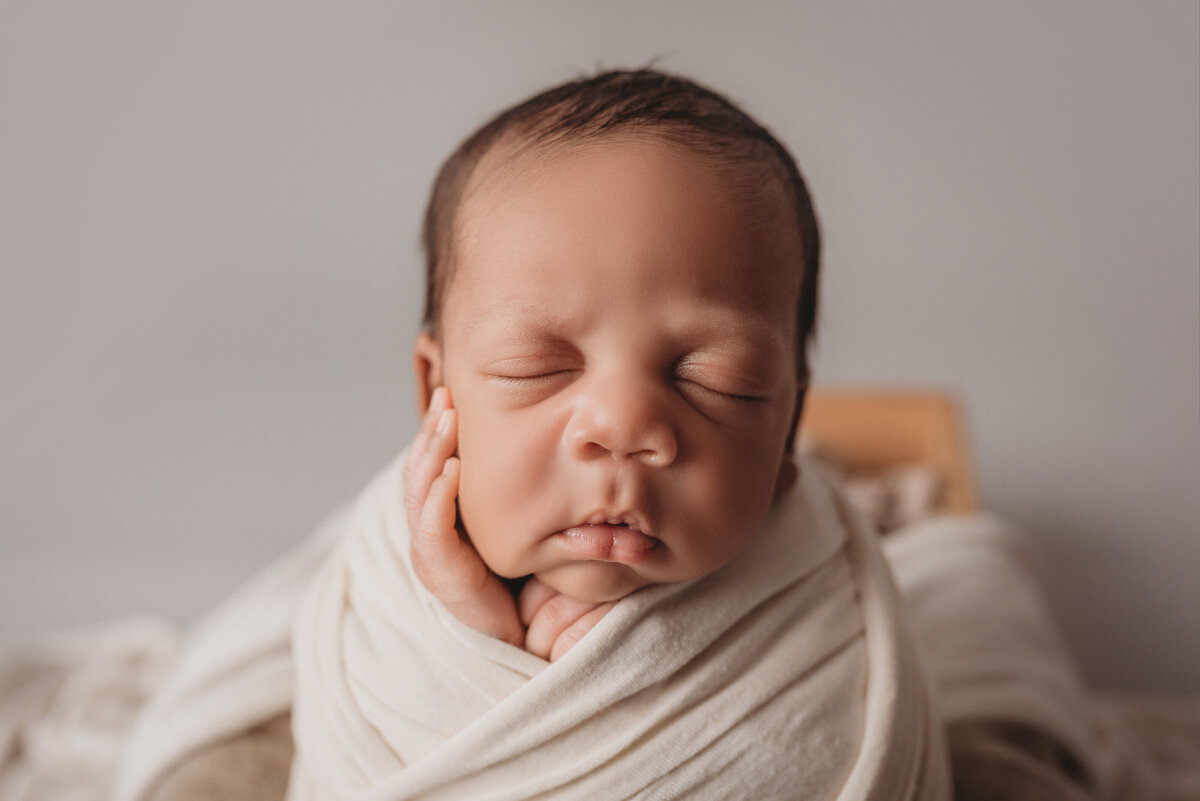 Newborn boy swaddled in white fabric sitting up with cheek in hand
