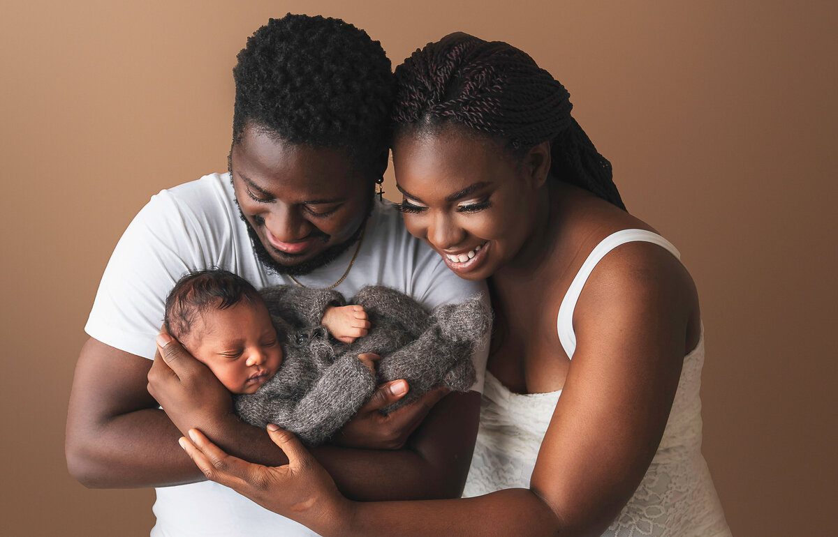 Dad holding baby boy in his arms facing the camera. Mom is standing behind dad's shoulder with her forearm touching dad's head. Mom and dad are looking at the sleeping baby boy and smiling at him.