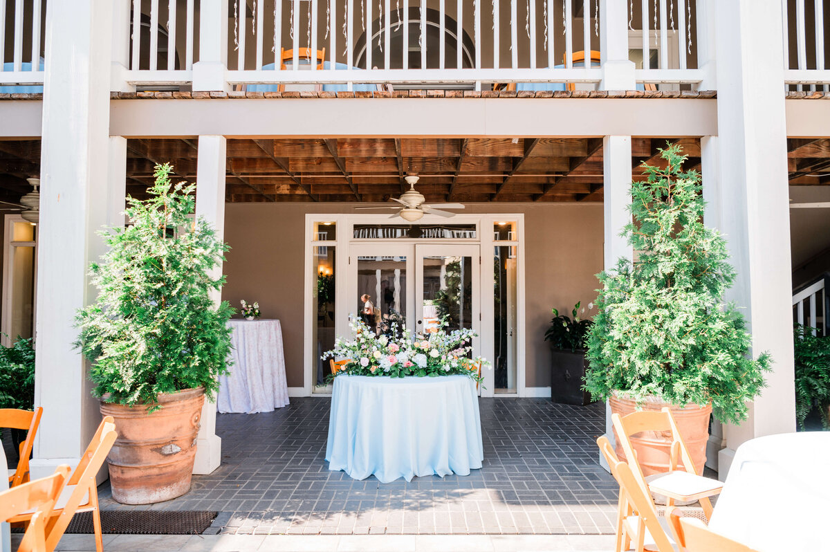 A round table with a light blue tablecloth and a floral centerpiece is set outside a building with large potted plants, wooden beams, and white railings. Chairs and more tables are visible around the area.