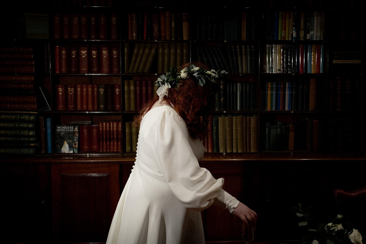 A bride walks past a book shelf in a library, her face covered by her red hair and flower crown
